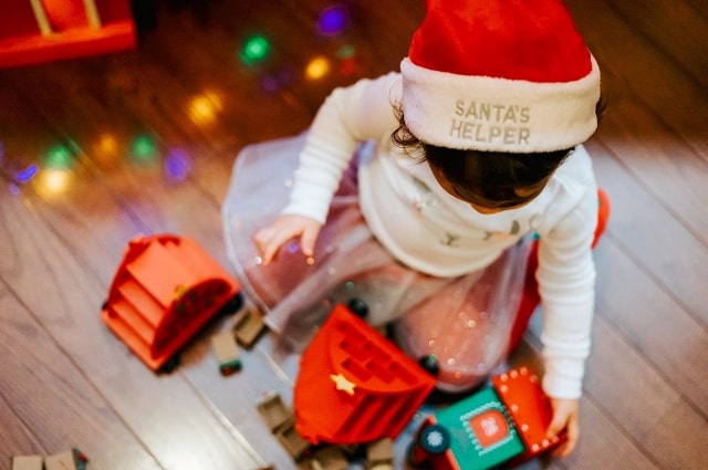 25 Under $25: Best Christmas Gifts for Baby, a baby girl wearing a Santa hat and playing on the floor with toys with holiday lights reflected on the wooden floor.